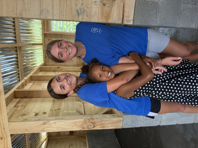 Two volunteers in blue shirts hugging a young local girl inside a wooden home